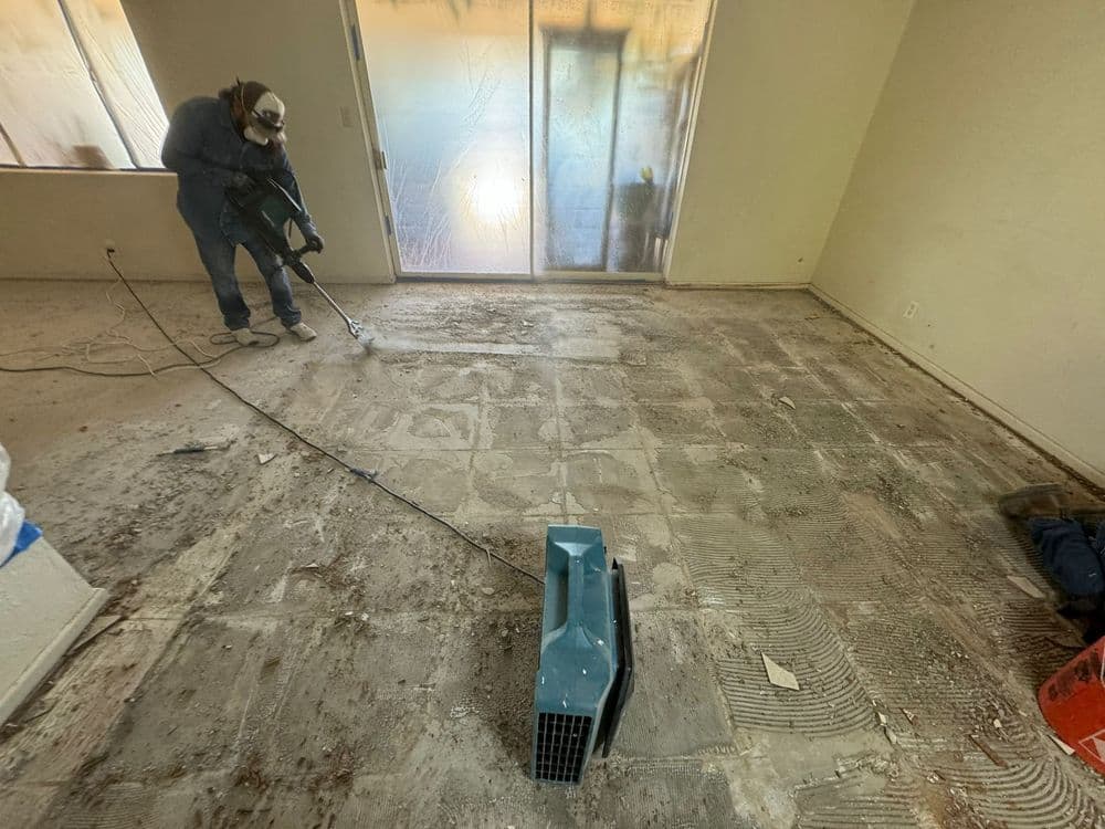 Worker using a floor scraper and air blower in an empty room during renovation.